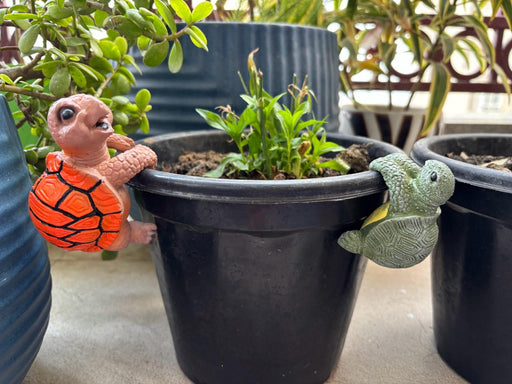 Two decorative turtle pot hangers, one orange and one green, climbing the rim of a black garden planter.
