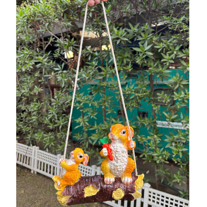 Whimsical hanging rabbit garden statue sitting on a rope swing and holding a carrot, outdoor resin bunny figurine for yard and patio decor.