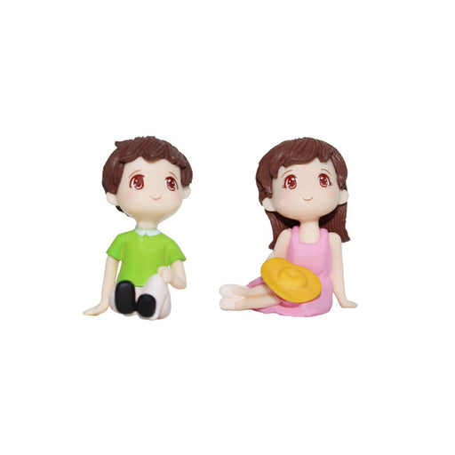 Miniature boy and girl figurines in a sitting pose, with the boy in a green shirt and the girl in a pink dress holding a yellow hat, isolated on a white background.