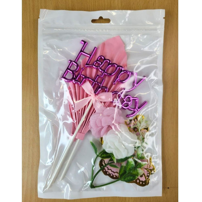 Close-up of a pink and gold butterfly cake topper set, including paper fan palm leaves, a Happy Birthday sign, and glitter butterflies, placed on a green grass background.