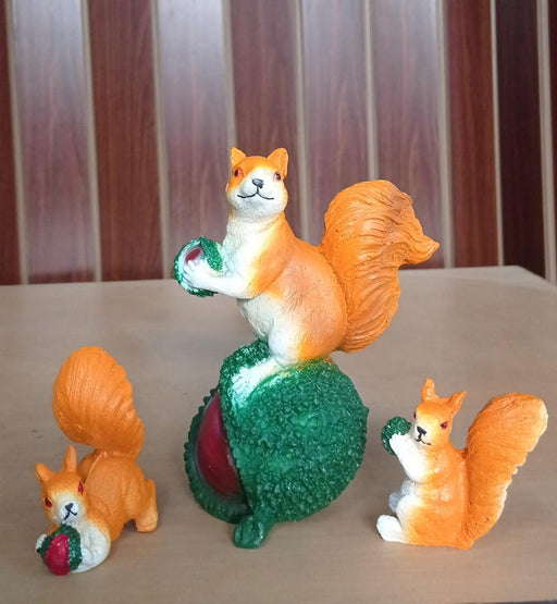 Three small, bright orange and white squirrel figurines holding red objects (likely nuts or berries). The central squirrel is perched on a large, textured green and red object (possibly a chestnut or pinecone), while the other two are seated on either side.