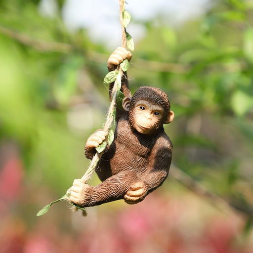 A realistic, hand-painted resin monkey figurine hanging from a rope vine with green leaves, positioned against a blurred outdoor garden background with green and pink foliage.
