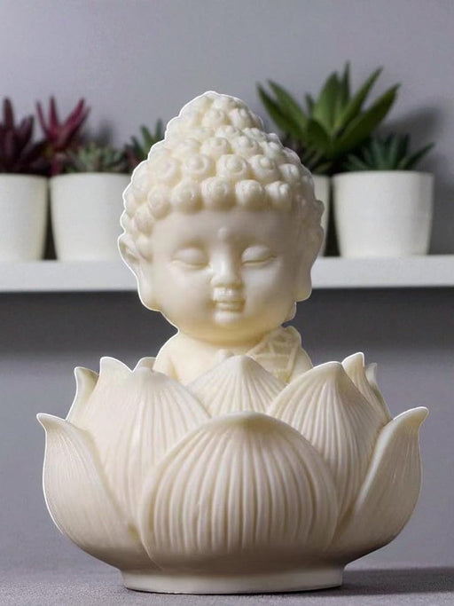 Small white resin Buddha statue resting in a blooming lotus flower base, with a backdrop of potted succulents on a shelf.