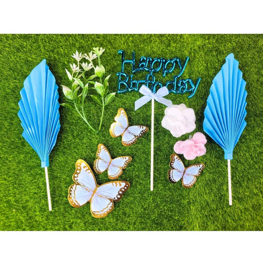 Close-up of a clear plastic package containing a Blue Butterfly and Happy Birthday cake topper set, featuring a large acrylic blue script and several blue feather accent picks.