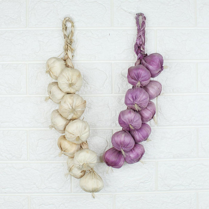 A pair of artificial hanging garlic braids hanging side-by-side against a white textured brick wall. The string on the left features off-white garlic bulbs, while the string on the right features purple garlic bulbs, both woven with rustic top loops.