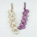 A pair of artificial hanging garlic braids hanging side-by-side against a white textured brick wall. The string on the left features off-white garlic bulbs, while the string on the right features purple garlic bulbs, both woven with rustic top loops.