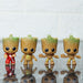 Four miniature Baby Groot figurines in different poses, including one in a red jumpsuit and three with natural wood textures, standing on a reflective surface.