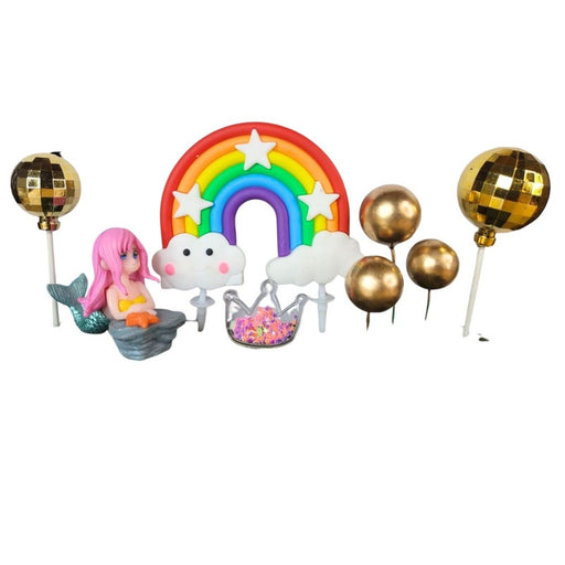 Close-up of a whimsical cake topper set featuring a pink-haired mermaid figurine, a large bright rainbow with white clouds, an iridescent crown pick, and three shiny gold sphere balloon toppers on sticks, all on a wooden surface.