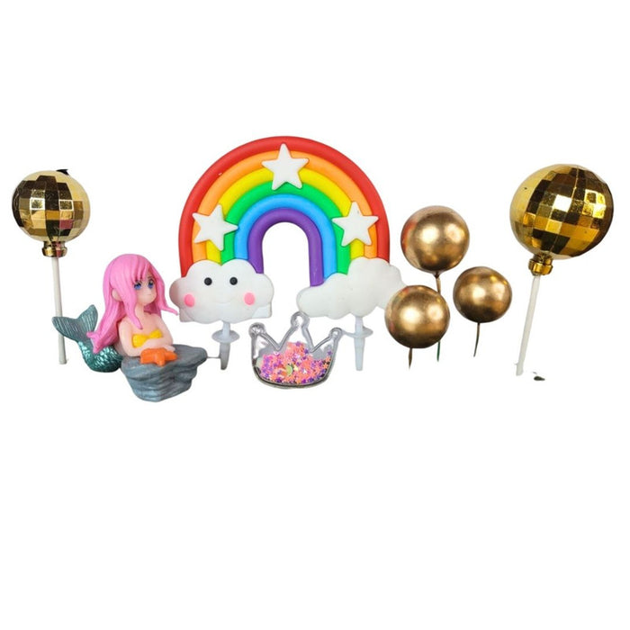 Close-up of a whimsical cake topper set featuring a pink-haired mermaid figurine, a large bright rainbow with white clouds, an iridescent crown pick, and three shiny gold sphere balloon toppers on sticks, all on a wooden surface.