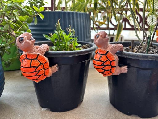 Two orange and tan decorative turtle pot hangers climbing on the rim of black garden planters with green plants.