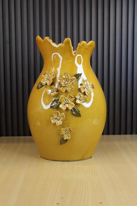 A decorative mustard yellow ceramic flower vase with a high-gloss finish, featuring a wavy scalloped rim and handcrafted 3D embossed yellow flowers with green leaves cascading down the front, isolated on a white background.