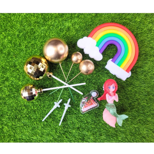 A complete set of cake toppers featuring a small, red-haired Mermaid figure, a colorful Rainbow with two white Clouds, a tiny banner, and several different-sized Metallic Gold Ball toppers on white and green picks, all displayed on a wooden table.