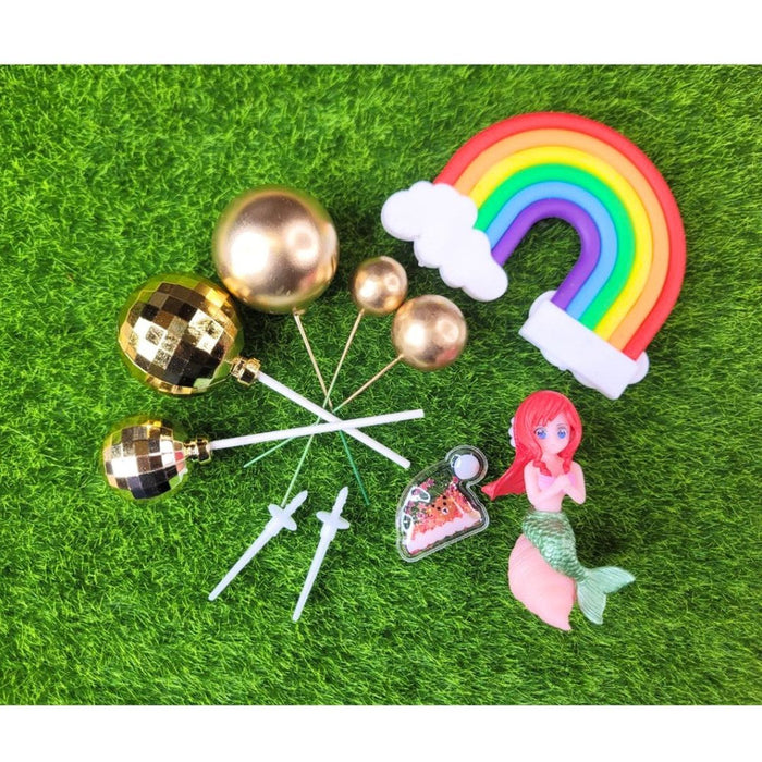 A complete set of cake toppers featuring a small, red-haired Mermaid figure, a colorful Rainbow with two white Clouds, a tiny banner, and several different-sized Metallic Gold Ball toppers on white and green picks, all displayed on a wooden table.