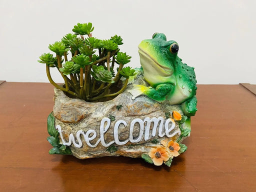 A decorative resin planter shaped like a rock with a green frog leaning on it and the word "welcome" sculpted across the front, holding small green succulent plants.