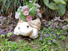 A cream-colored turtle-shaped garden planter filled with pink and white flowers, sitting on soil surrounded by green and purple garden plants.