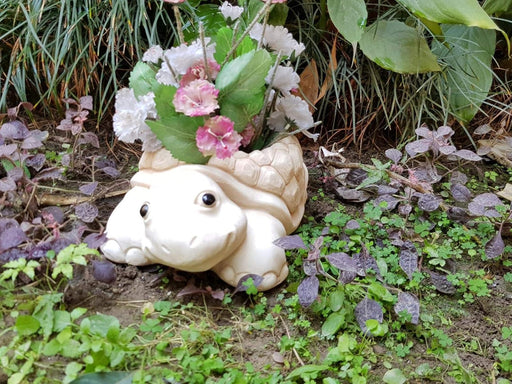 A cream-colored turtle-shaped garden planter filled with pink and white flowers, sitting on soil surrounded by green and purple garden plants.