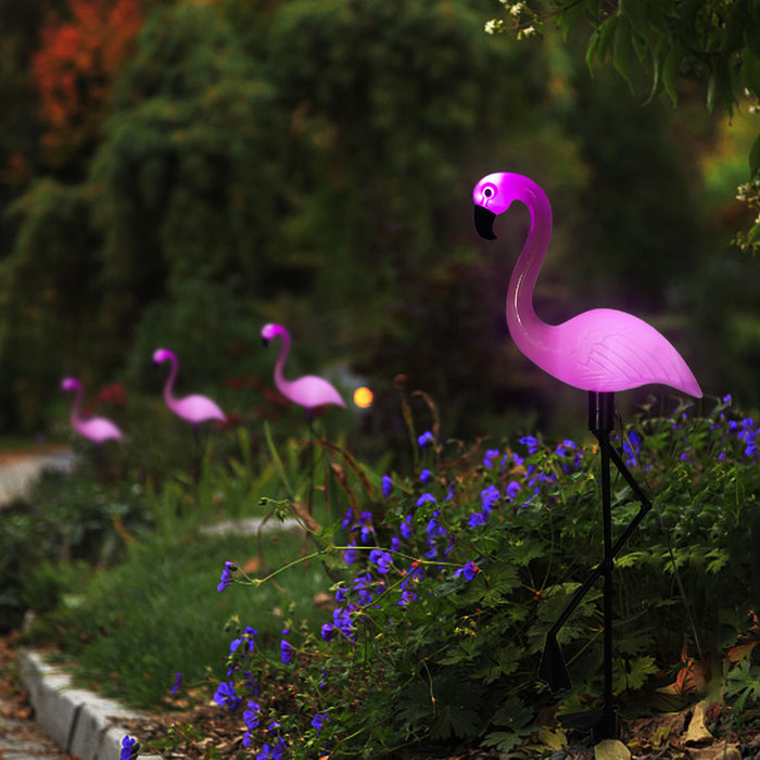 "A set of four pink plastic flamingo solar garden lights with black metal stakes standing in a lush green garden with rocks and white flowers."