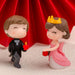 Miniature romantic couple figurines featuring a groom in a black tuxedo and a bride in a pink dress and golden crown, set against a red background.