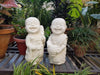 Two white laughing little monk statues sitting among potted plants in a garden setting.