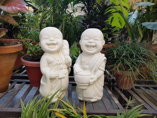 Two white laughing little monk statues sitting among potted plants in a garden setting.