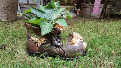 A detailed resin garden planter shaped like a weathered old boot with miniature rabbits peeking out and a green plant growing from the top, set on grass.