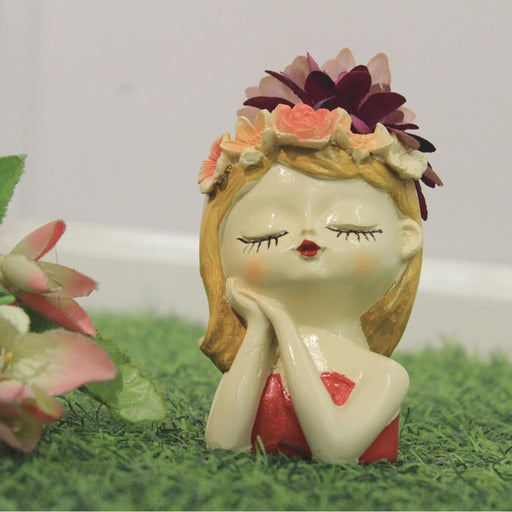 A decorative resin planter shaped like a girl's head with closed eyes and a floral crown, resting on her hands. The planter contains dark purple flowers and is shown on green artificial grass.