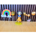 Close-up of a plastic-wrapped Snow White-style doll cake topper set including a dark-haired doll figure in a blue and orange dress, a colorful rainbow slice, a white cloud wand, and several shiny metallic gold ball ornaments, all on a wooden surface.