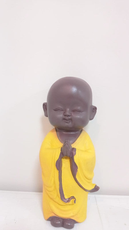 Buddhist monk statue in a bright yellow robe, standing with hands pressed together at the chest against a light background.