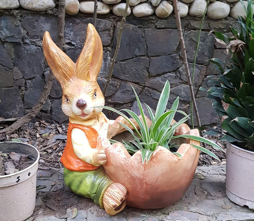 A decorative garden planter featuring a rabbit figurine wearing an orange vest and green pants, sitting and holding a large, cracked eggshell-shaped bowl that contains a small green plant. The statue is placed outdoors against a stone wall.