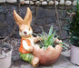 A decorative garden planter featuring a rabbit figurine wearing an orange vest and green pants, sitting and holding a large, cracked eggshell-shaped bowl that contains a small green plant. The statue is placed outdoors against a stone wall.