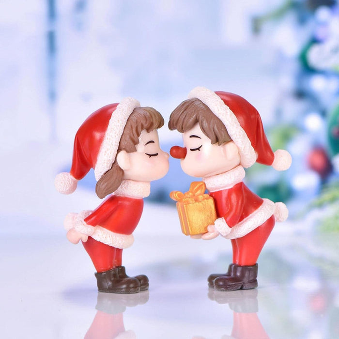Close-up of two small resin miniature figurines: a boy and a girl, both wearing red and white Santa outfits. The boy has a red nose and is holding a small golden gift box toward the girl. 
