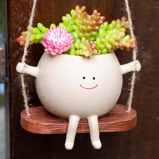 A cute round ceramic planter with a smiley face sitting on a miniature wooden swing, holding green succulent plants and hanging by jute ropes against a soft green background.