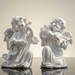 Two white cherub angel figurines kneeling and playing musical instruments against a dark outdoor background.