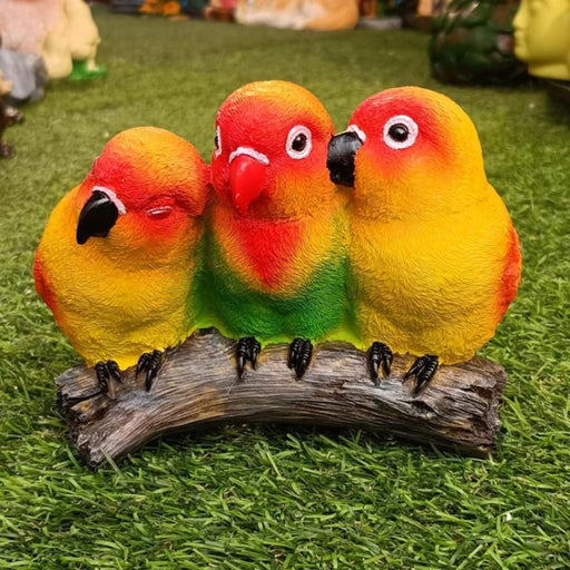 Three colorful lovebird figurines, painted red, yellow, and green, sitting close together on a faux log, resting on a bright green artificial turf.