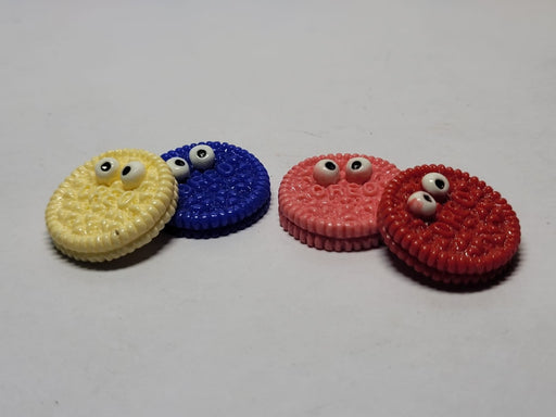 Four colorful novelty cookie-shaped charms or cabochons—one yellow, one blue, one pink, and one red—each decorated with small googly eyes, sitting on a white background.