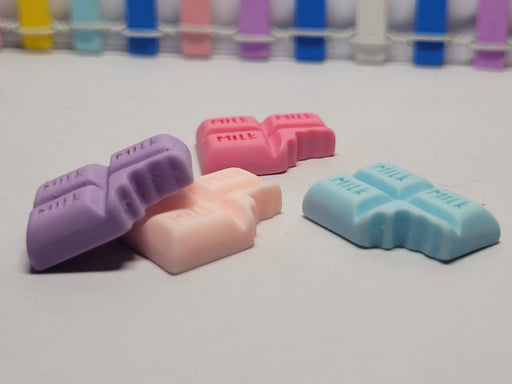 Four miniature chocolate bar-shaped cabochons or wax melts in pastel colors: purple, pink, light pink (or peach), and light blue, each with the word 'MILK' embossed on the squares