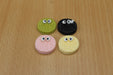 Four resin cookie-shaped charms with googly eyes, colored green, black, pink, and yellow, arranged in a square on a light brown wooden surface.