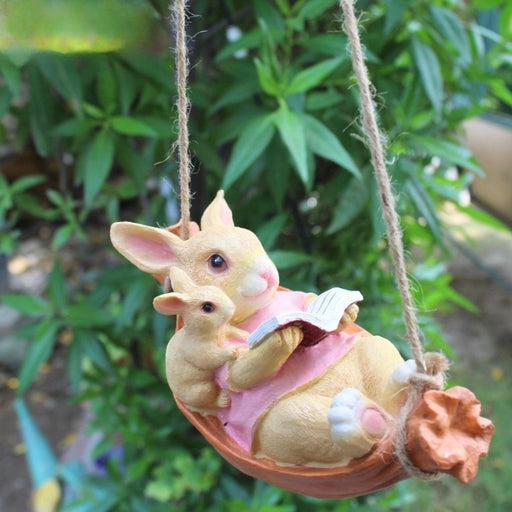 A brown rabbit figurine wearing a pink dress, lying in a carrot-shaped swing and reading a book with a small mouse beside it.