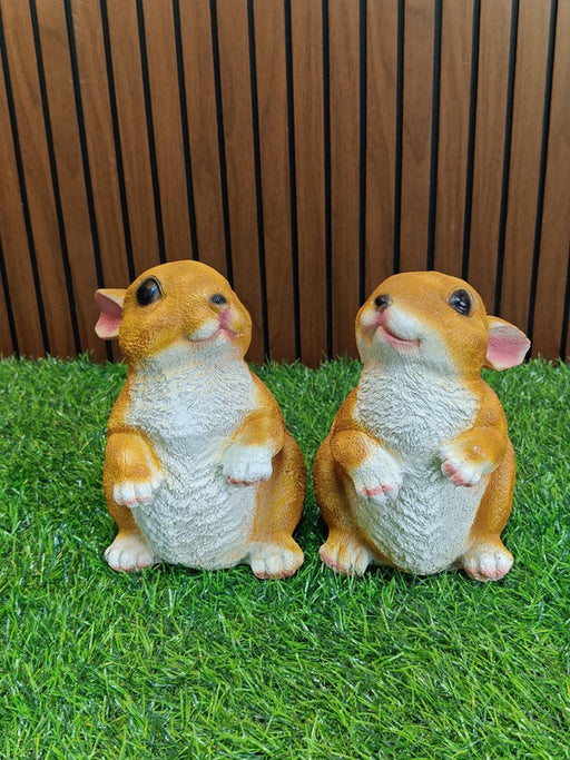 Two identical, cute, small decorative resin figurines of baby rabbits or hamsters sitting upright on bright green artificial grass, set against a background of dark vertical wooden panels.