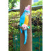 Colorful resin parrot garden decoration (blue, yellow, pink) mounted on a brown tree in a green outdoor setting.