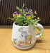 A rustic ceramic planter shaped like a large cup featuring a vintage illustration of blue tit birds, a blue birdhouse, and spring flowers on a weathered white background with an attached saucer.