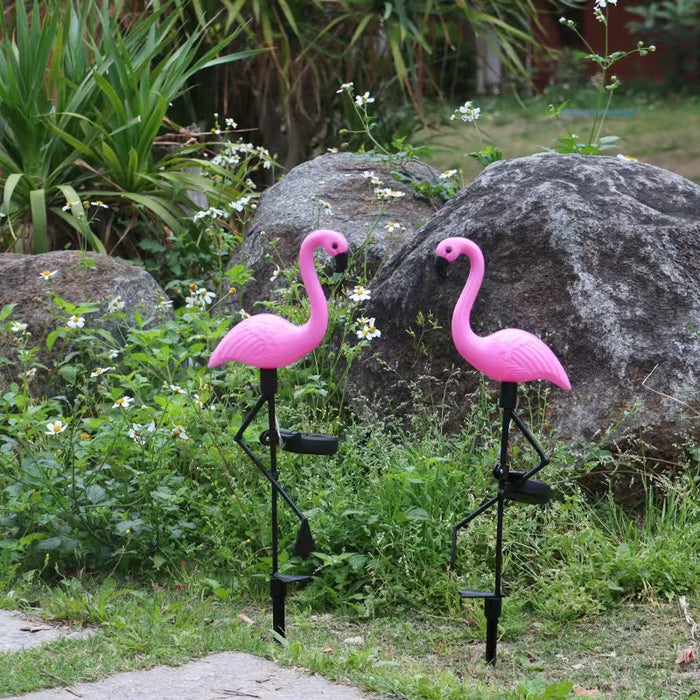 "A set of two pink plastic flamingo solar garden lights with black metal stakes standing in a lush green garden with rocks and white flowers."
