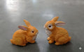 Two brown ceramic rabbit figurines with pink ears and red eyes sitting on a grey floor.