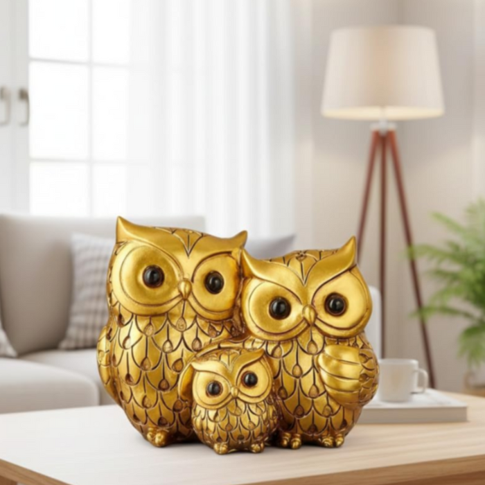 Three golden owl figurines of varying sizes—representing a family—displayed on a wooden coffee table in a brightly lit, modern living room with a blurred sofa and floor lamp in the background.
