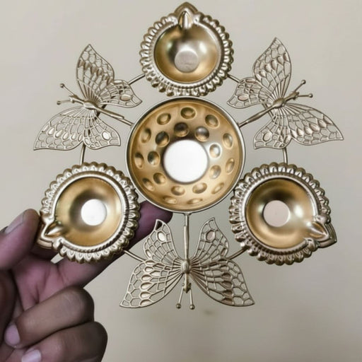 A hand holds a decorative gold-colored metal diya holder designed with four intricate filigree butterflies. The stand features a central bowl surrounded by four traditional oil lamps (diyas), creating a five-wick centerpiece for festive home decor.