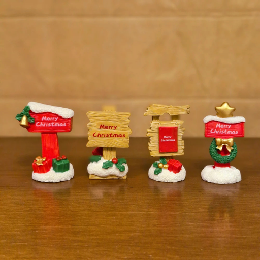 A set of four brightly colored miniature resin Christmas signs, each on a snowy base, reading "Merry Christmas." The signs feature details like a bell, presents, a wooden texture, and a gold star with a wreath.
