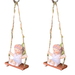 Two identical whimsical resin figurines of a young girl with a flower crown sitting on a wooden-style swing, suspended by twine ropes decorated with green fabric leaves.