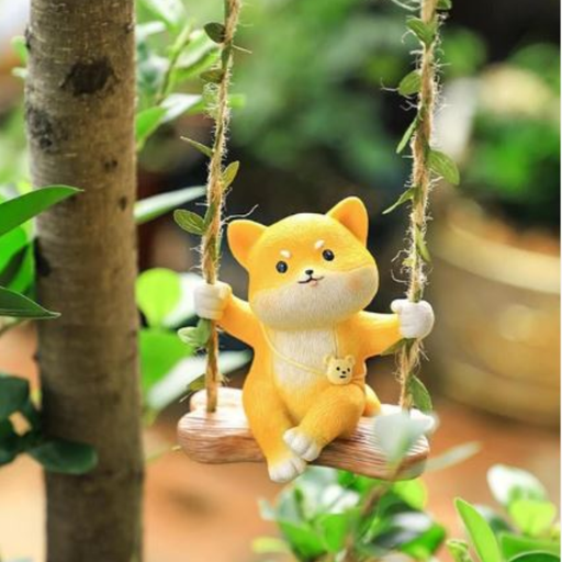 A miniature garden figurine of a ginger kitten sitting on a small rope swing suspended from a tree branch, surrounded by green foliage.