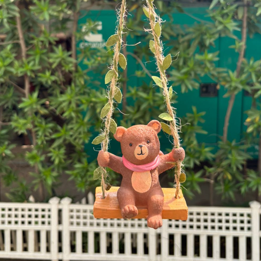 A garden statue of a brown teddy bear sitting on a wooden swing suspended by rope-textured chains, positioned in front of a white picket fence and green foliage.