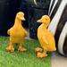 Two identical light brown duckling figurines standing side-by-side on a light surface with a blurred outdoor background.